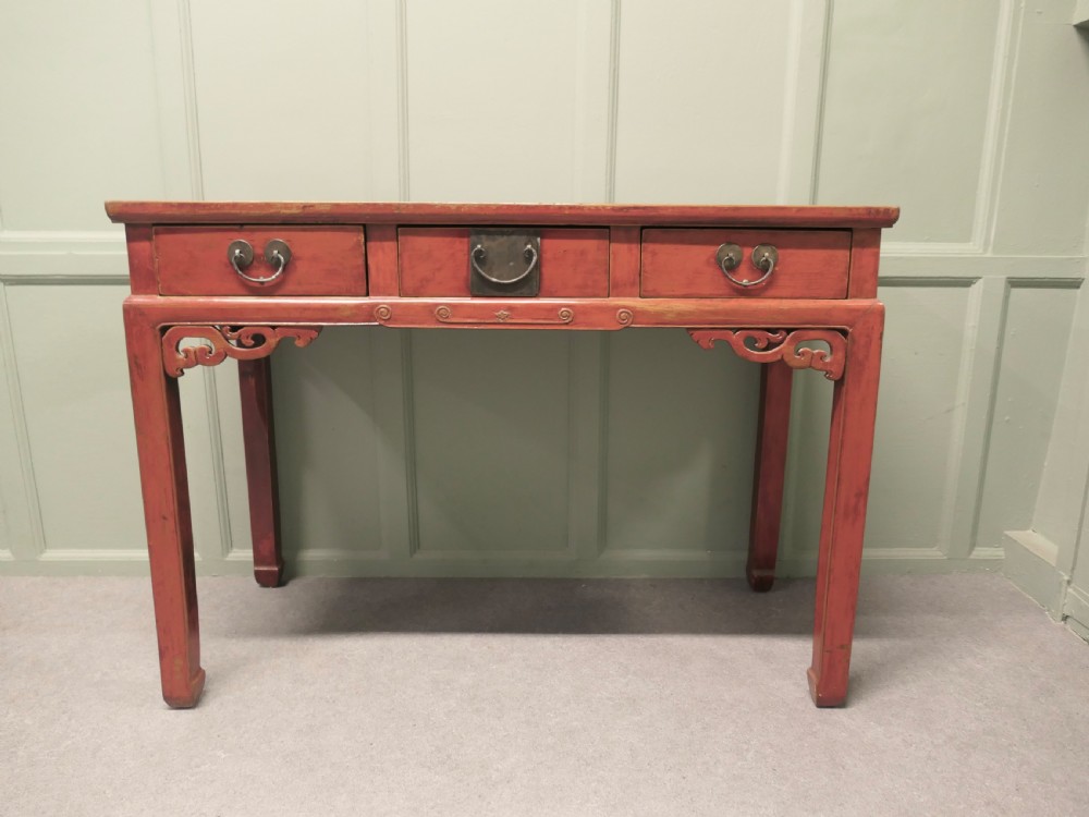 A red lacquered timber console table featuring three drawers with brass drop handles and carved corner spandrels over straight square legs.