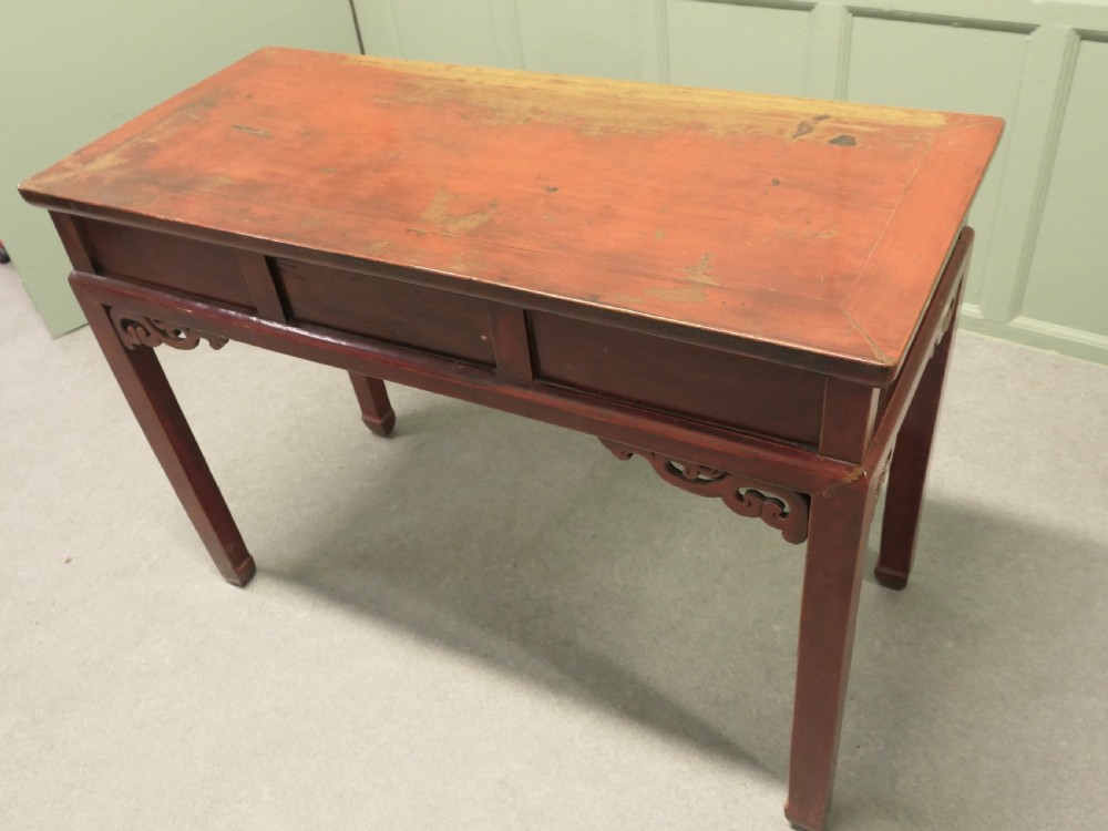 A red lacquered timber console table featuring three drawers with brass drop handles and carved corner spandrels over straight square legs. View V