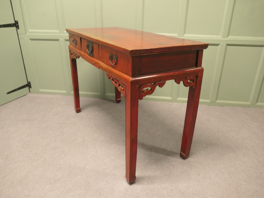 A red lacquered timber console table featuring three drawers with brass drop handles and carved corner spandrels over straight square legs. View VI
