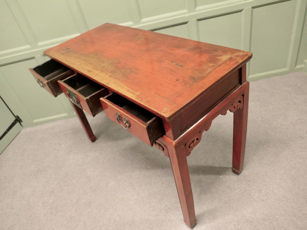 A red lacquered timber console table featuring three drawers with brass drop handles and carved corner spandrels over straight square legs. View VII