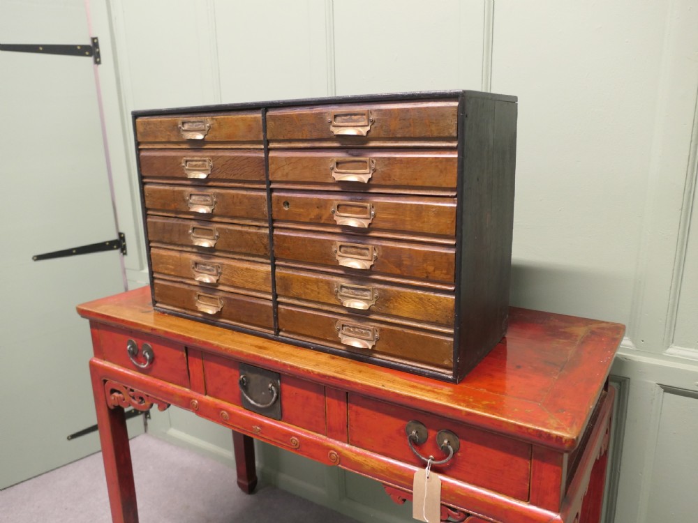 A desktop oak cabinet featuring twelve shallow drawers arranged in two columns, fitted with brass cup handles and label holders.