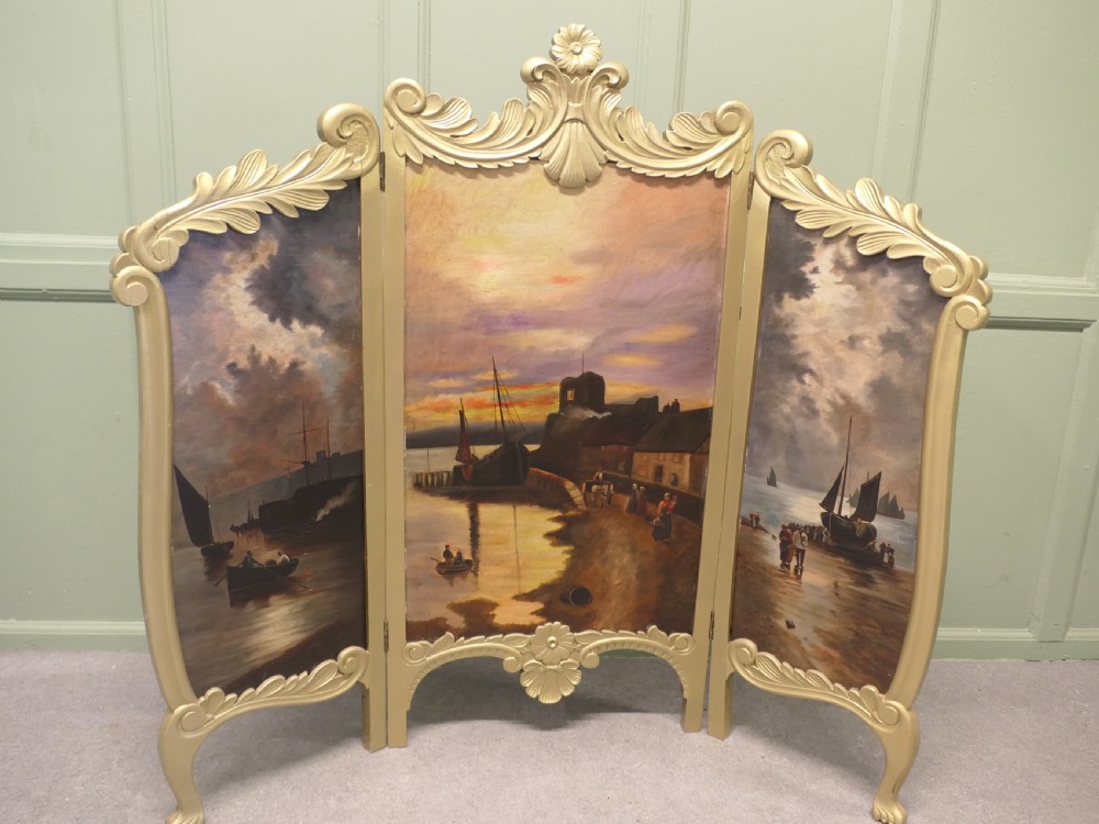A three-panel folding fire screen with a carved gold giltwood frame and oil-painted maritime scenes depicting ships near a coastal shore at sunset.