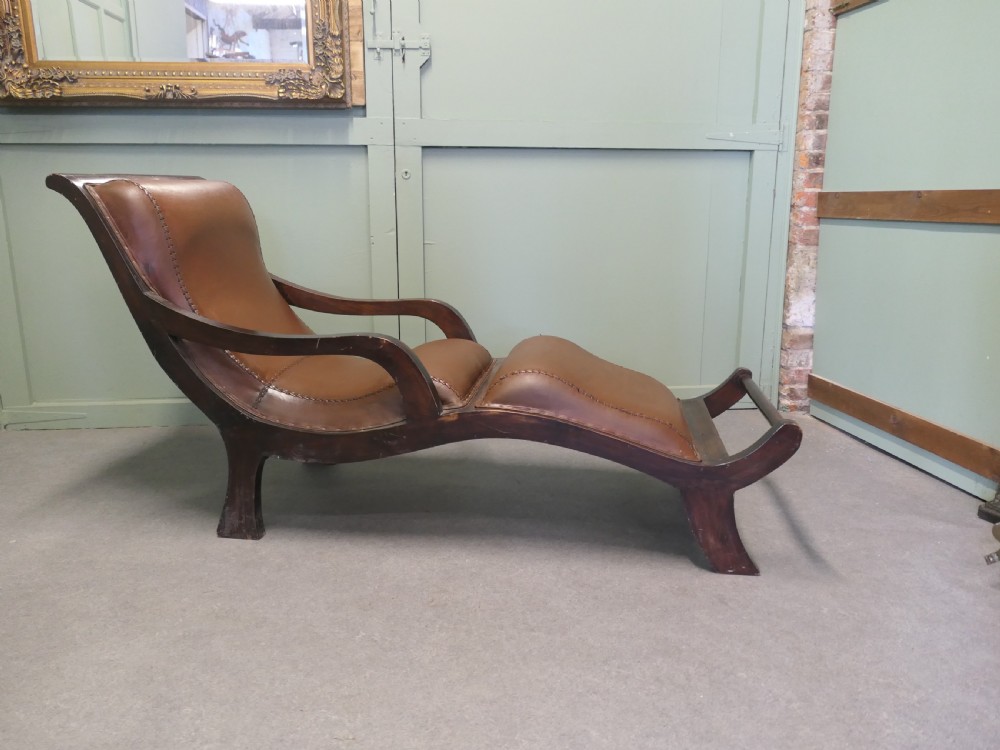 A Victorian colonial plantation chaise longue featuring a curved mahogany frame, extended arms, and rich brown leather upholstery with metal stud detailing.