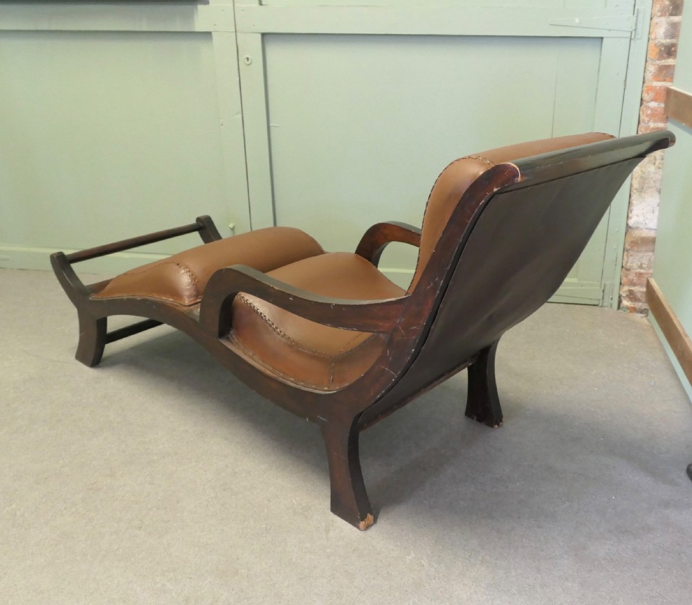 A Victorian colonial plantation chaise longue featuring a curved mahogany frame, extended arms, and rich brown leather upholstery with metal stud detailing. View V