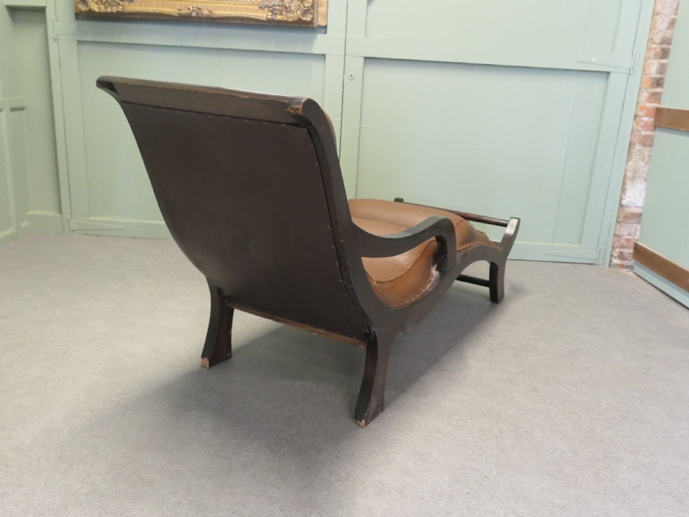 A Victorian colonial plantation chaise longue featuring a curved mahogany frame, extended arms, and rich brown leather upholstery with metal stud detailing. View VI
