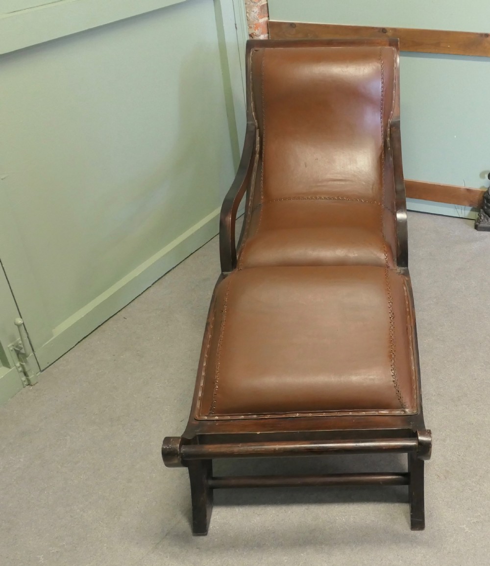 A Victorian colonial plantation chaise longue featuring a curved mahogany frame, extended arms, and rich brown leather upholstery with metal stud detailing. View VII