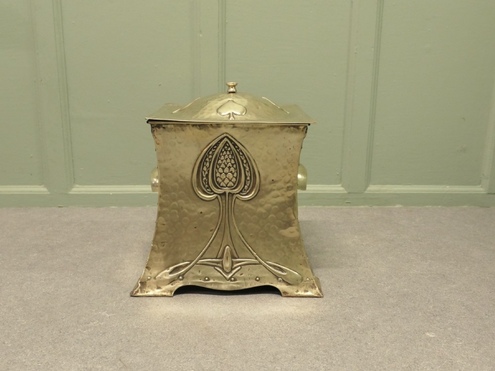 An antique hammered brass coal box featuring a stylised plant motif embossed on the front panel and a hinged top lid.