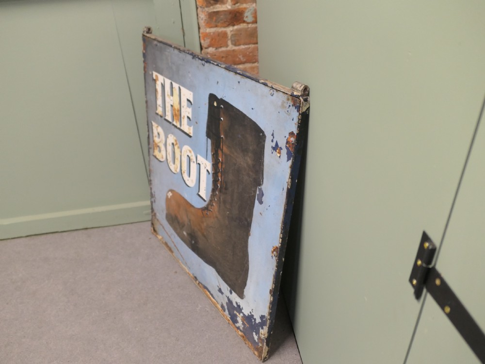 A square weathered painted iron pub sign depicting a brown boot alongside white text reading THE BOOT against a light blue background. View V