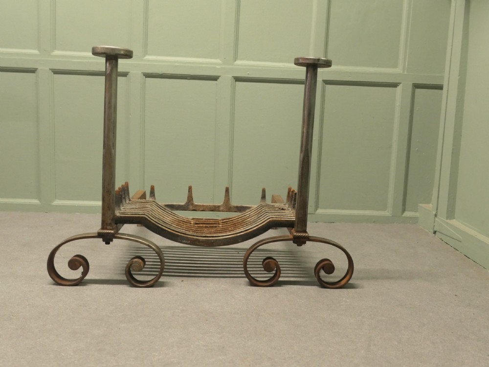 A heavy dark iron fireplace grate featuring scrolled front feet, twin upright pillar posts with flat circular tops, and a curved central log basket.