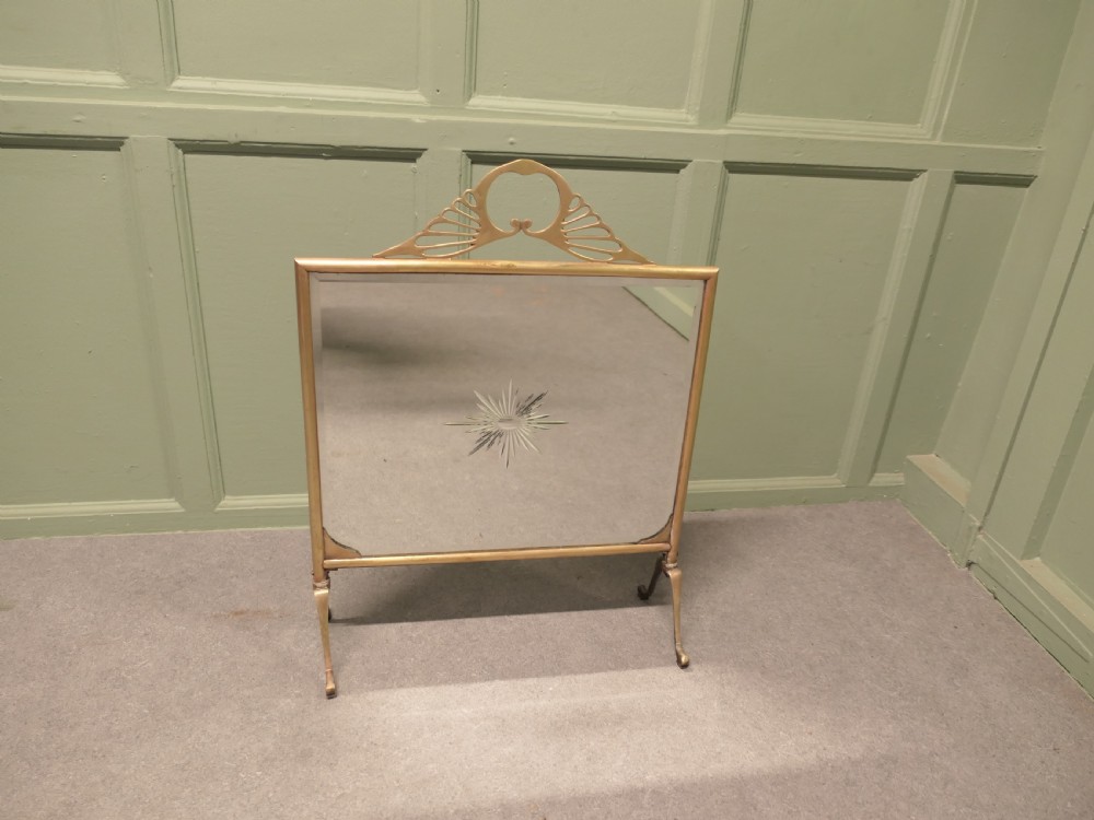 An Art Deco brass fire screen featuring a mirrored glass panel with a central etched sunburst motif and splayed feet.