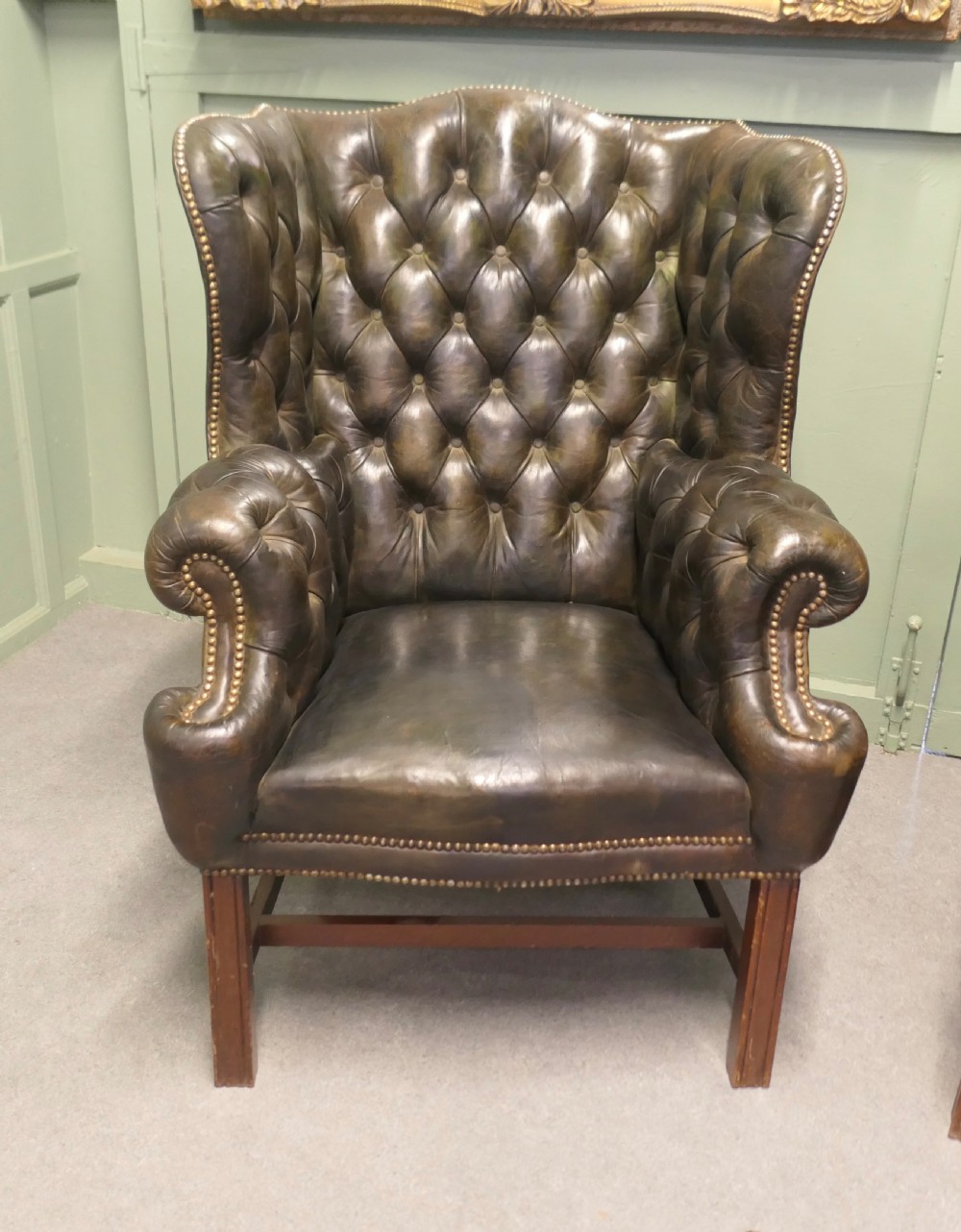 A pair of olive green leather wingback armchairs featuring deep button tufting, brass studding, and square wooden legs. View III