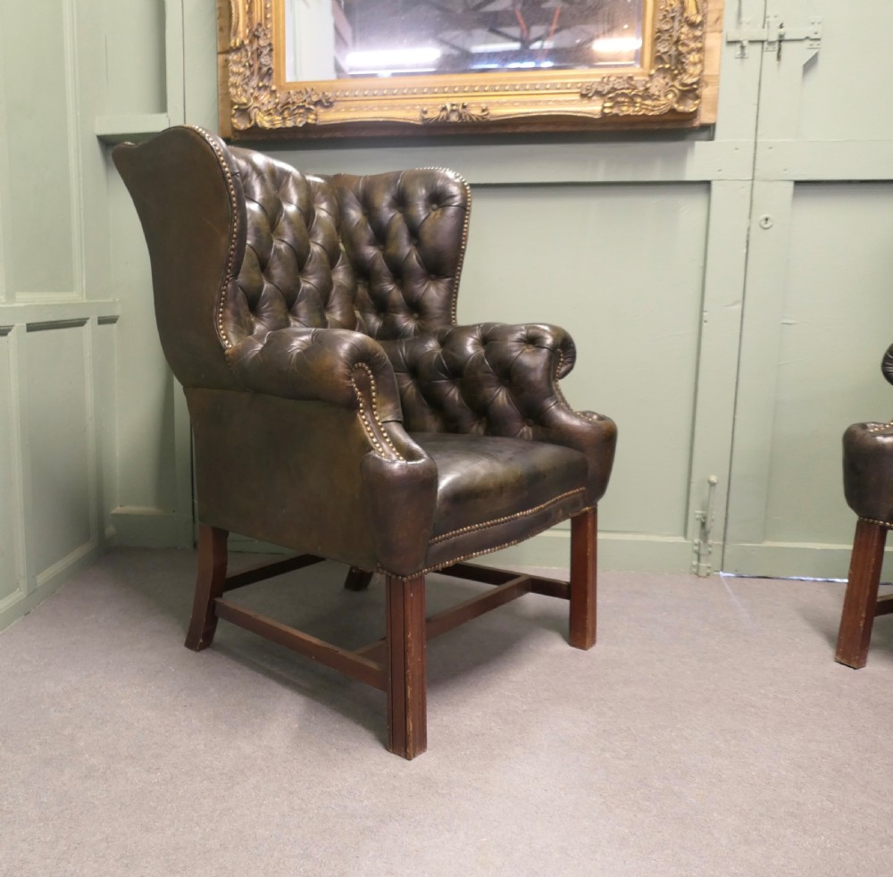 A pair of olive green leather wingback armchairs featuring deep button tufting, brass studding, and square wooden legs. View V