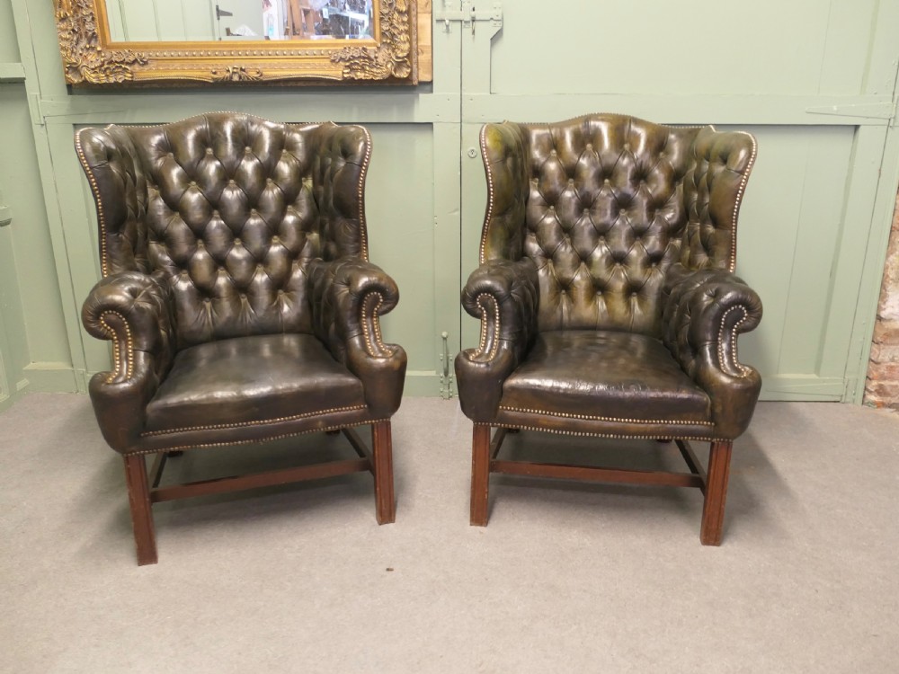 A pair of olive green leather wingback armchairs featuring deep button tufting, brass studding, and square wooden legs. View VI