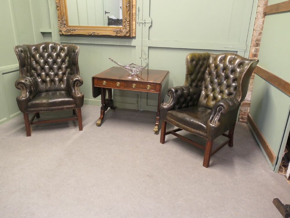 A pair of olive green leather wingback armchairs featuring deep button tufting, brass studding, and square wooden legs. View VII
