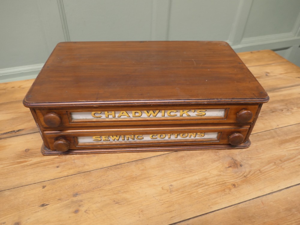 A two-drawer timber haberdashery display cabinet with turned wooden knobs and glass drawer fronts featuring gold lettering that reads Chadwicks Sewing Cottons.