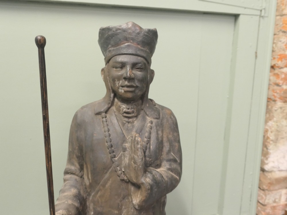 A dark terracotta sculpture depicting a standing priest figure wearing a hat and prayer beads while holding a wooden staff.