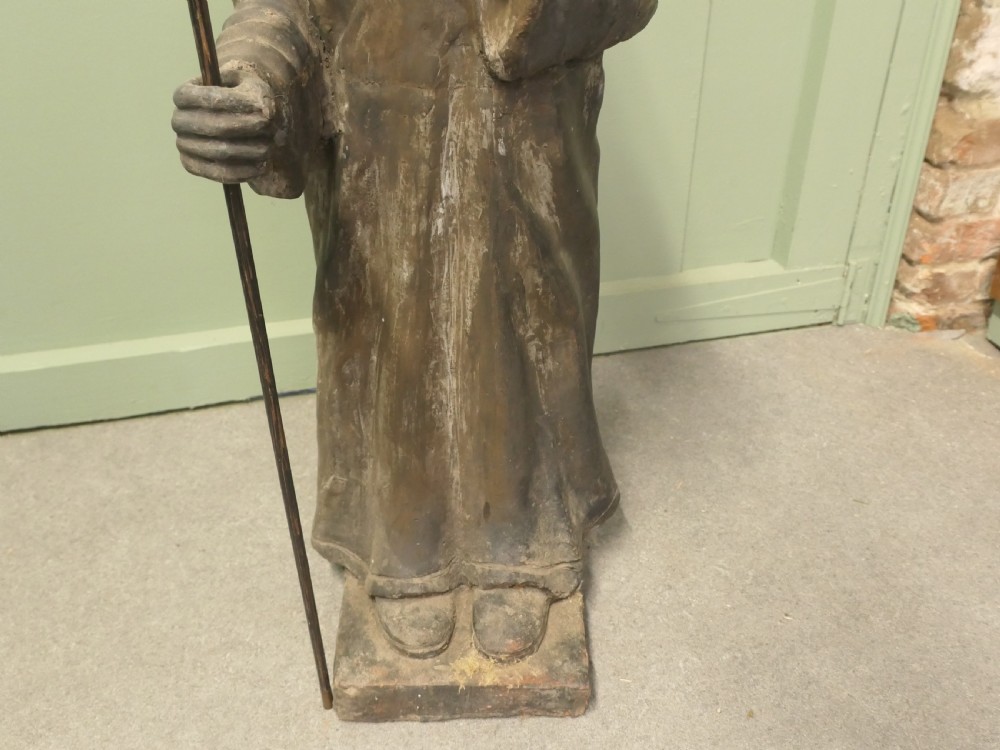 A dark terracotta sculpture depicting a standing priest figure wearing a hat and prayer beads while holding a wooden staff. View IV