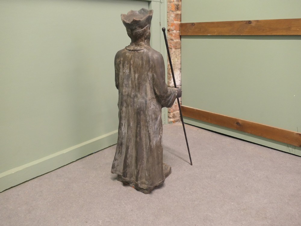 A dark terracotta sculpture depicting a standing priest figure wearing a hat and prayer beads while holding a wooden staff. View V