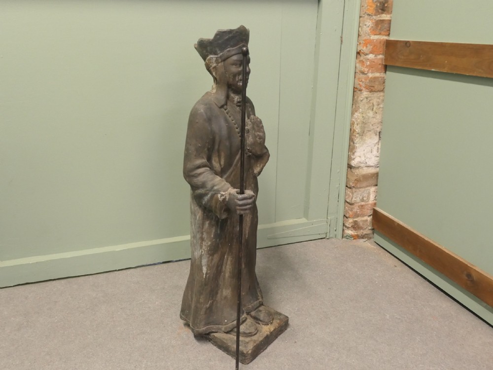 A dark terracotta sculpture depicting a standing priest figure wearing a hat and prayer beads while holding a wooden staff. View VI