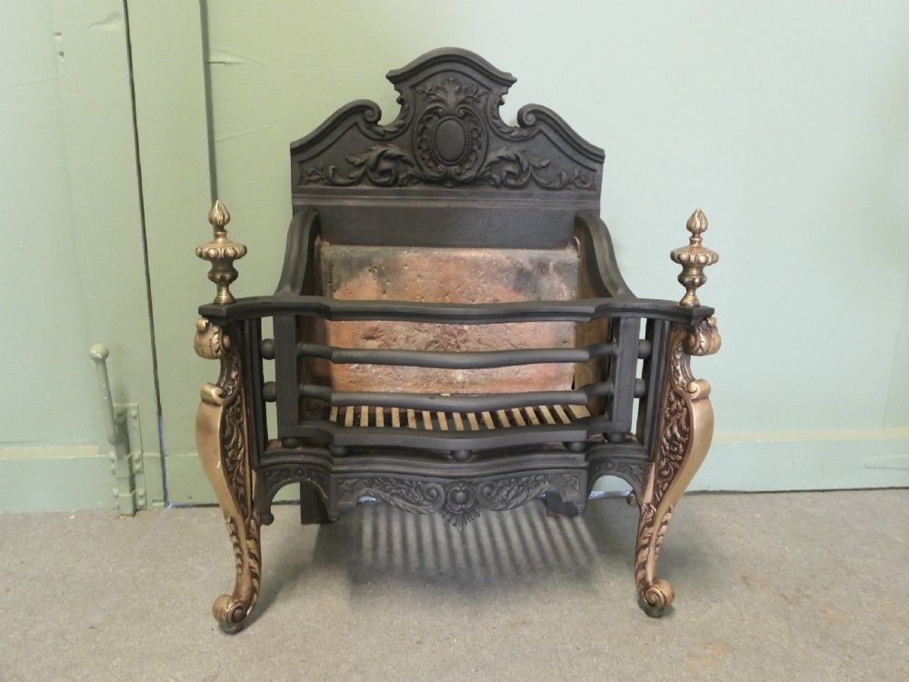 A freestanding cast iron fire basket with brass scroll legs, urn finials, an ornate crest backplate, and an internal firebrick lining.