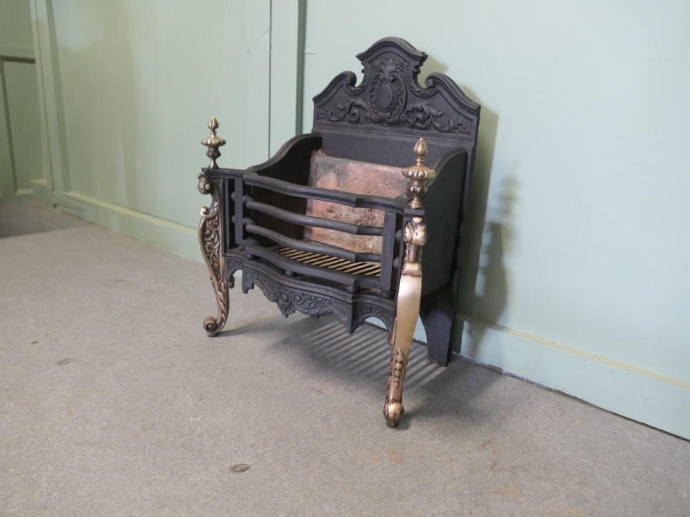 A freestanding cast iron fire basket with brass scroll legs, urn finials, an ornate crest backplate, and an internal firebrick lining. View II