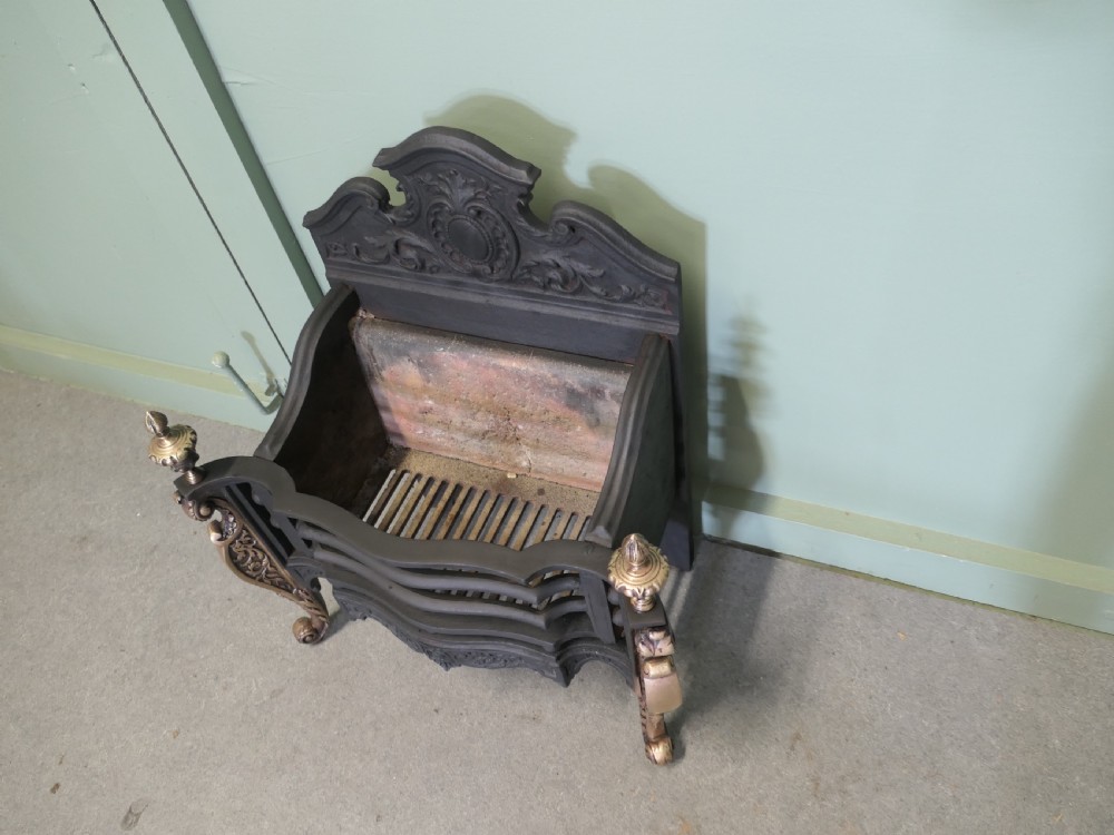 A freestanding cast iron fire basket with brass scroll legs, urn finials, an ornate crest backplate, and an internal firebrick lining. View V