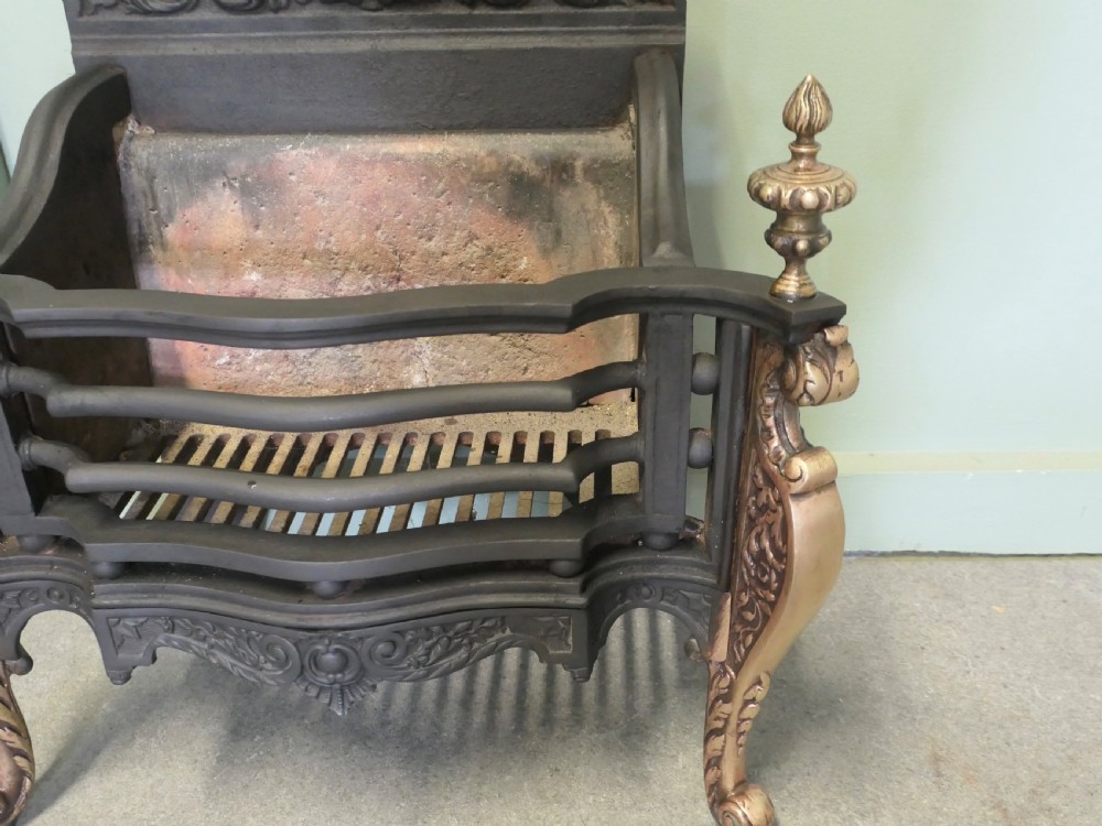 A freestanding cast iron fire basket with brass scroll legs, urn finials, an ornate crest backplate, and an internal firebrick lining. View VI