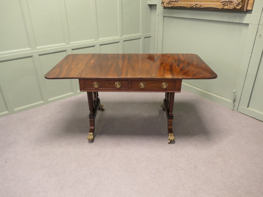 A Regency mahogany sofa table featuring two drop leaves, dual frieze drawers with brass handles, and lyre-shaped end supports terminating in brass lion paw castors.