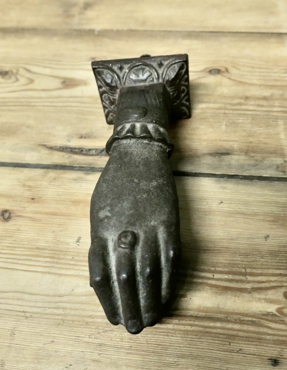 A cast iron door knocker sculpted in the shape of a hand with a detailed frilled cuff attached to a decorated square backplate.