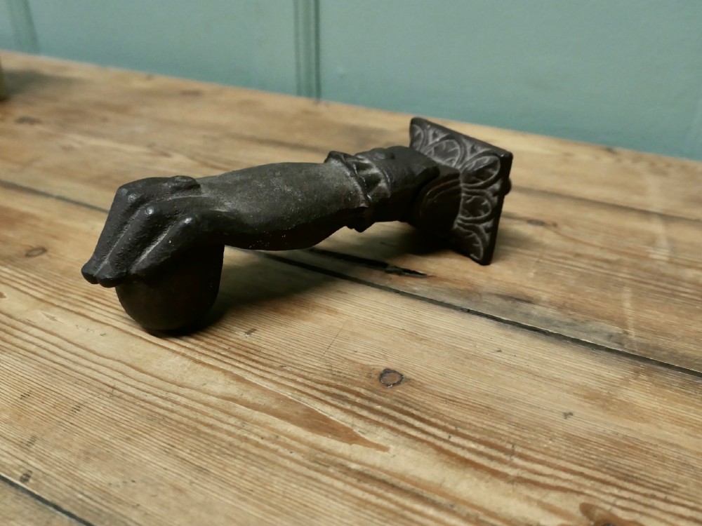 A cast iron door knocker sculpted in the shape of a hand with a detailed frilled cuff attached to a decorated square backplate. View II