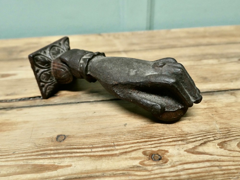 A cast iron door knocker sculpted in the shape of a hand with a detailed frilled cuff attached to a decorated square backplate. View III