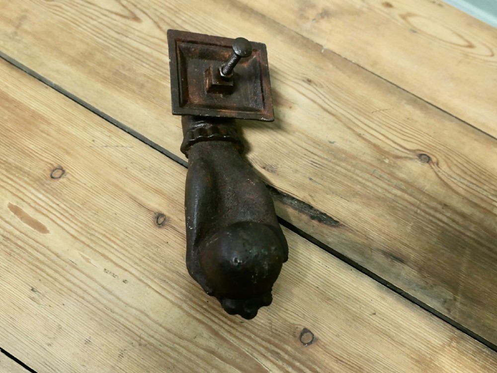 A cast iron door knocker sculpted in the shape of a hand with a detailed frilled cuff attached to a decorated square backplate. View IV