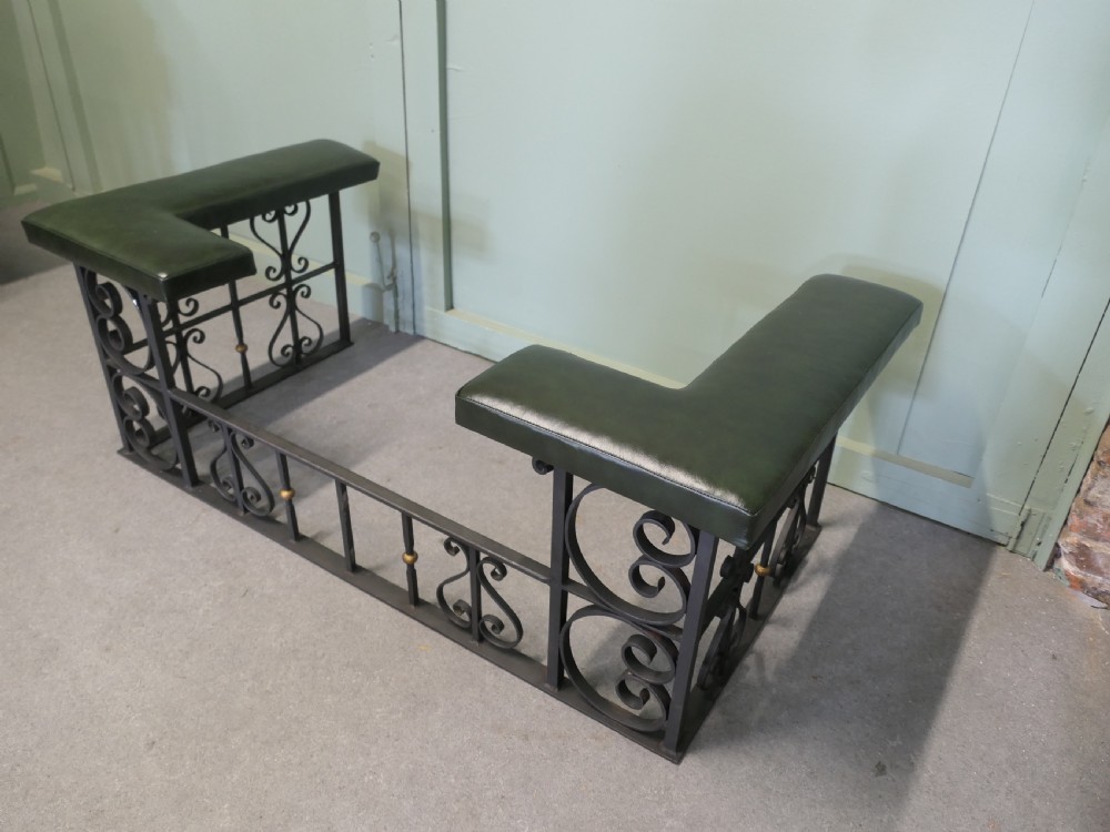 A black wrought iron club fender featuring scrollwork framing, brass details, and two green leather L-shaped corner cushions.