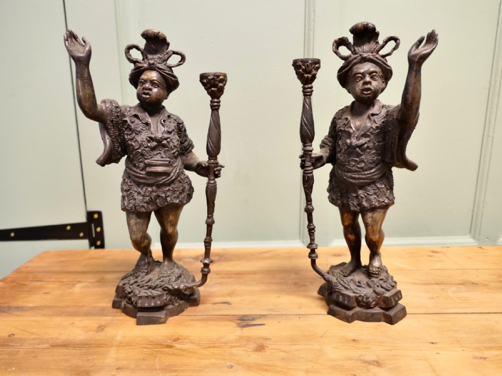 A pair of dark patinated cast brass figural candlesticks depicting standing figures in patterned tunics and turbans holding raised candle nozzles.