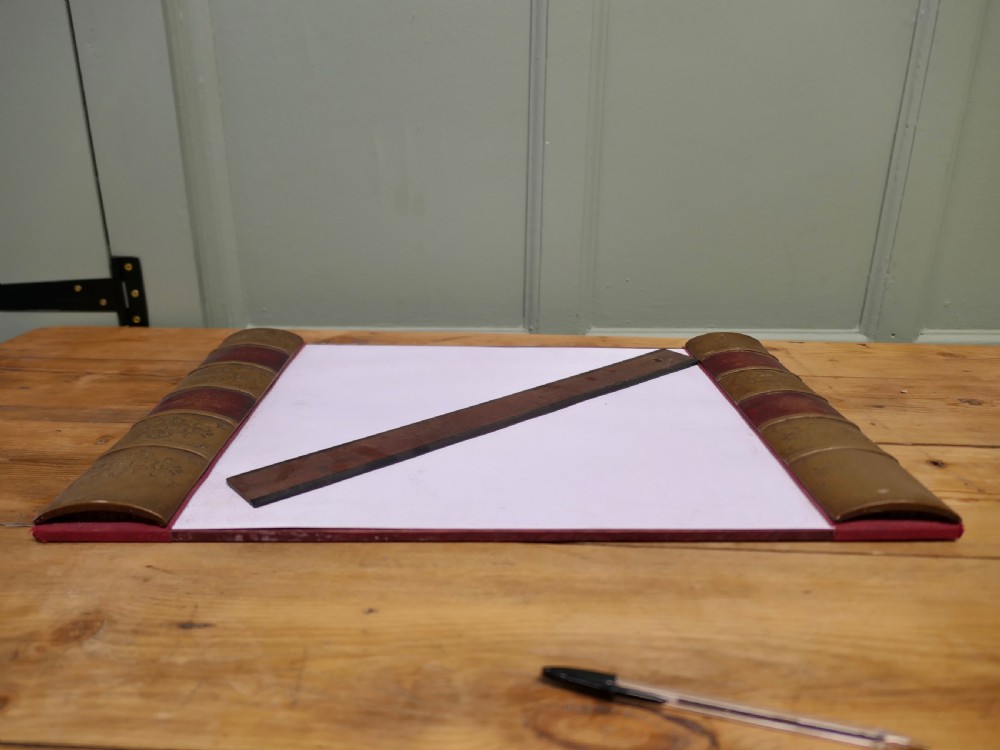 An antique leather desk blotter featuring faux book spine side borders with gold leaf tooling and a wooden ruler lying across the central pad. View II
