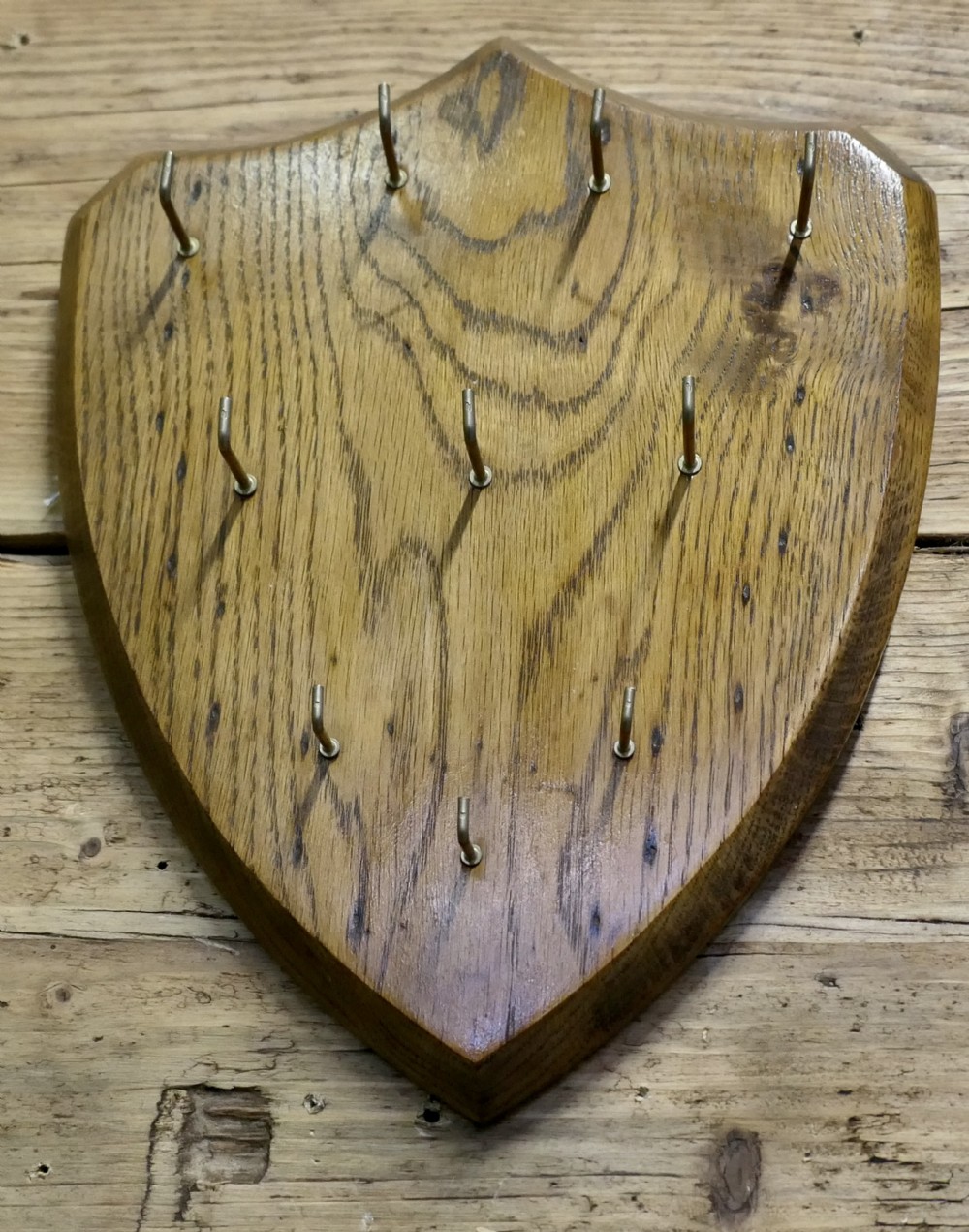 A shield-shaped solid oak board featuring eleven brass L-hooks arranged systematically across its polished grain surface.