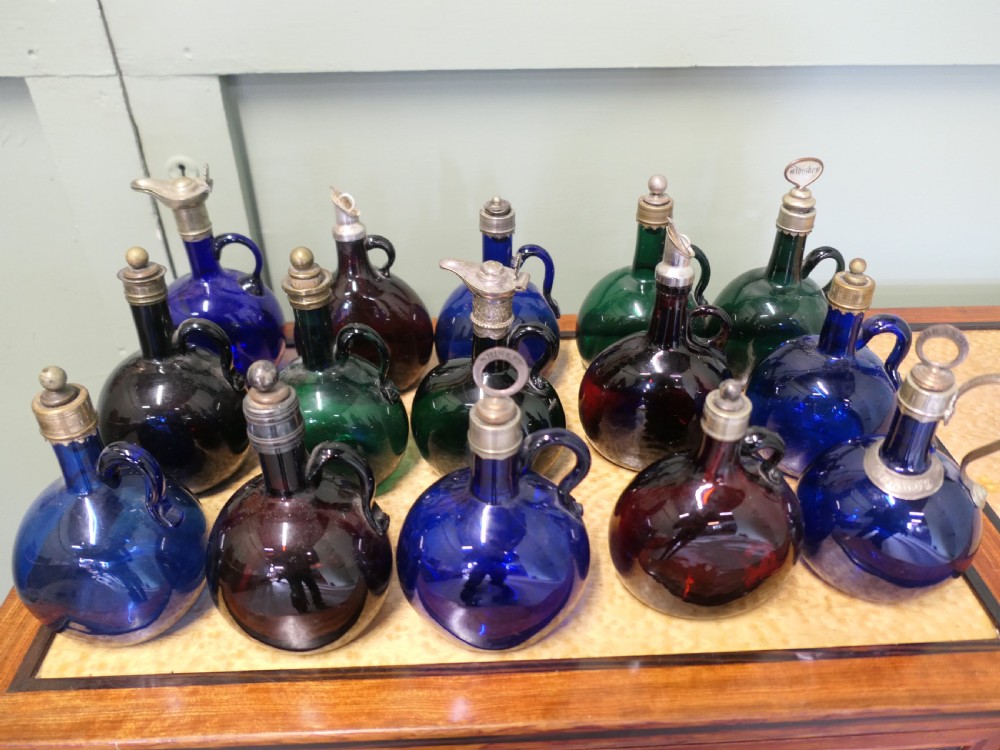 A collection of fifteen 19th-century Victorian coloured glass flagon decanters with metal spouts and stoppers arranged on a wooden surface.