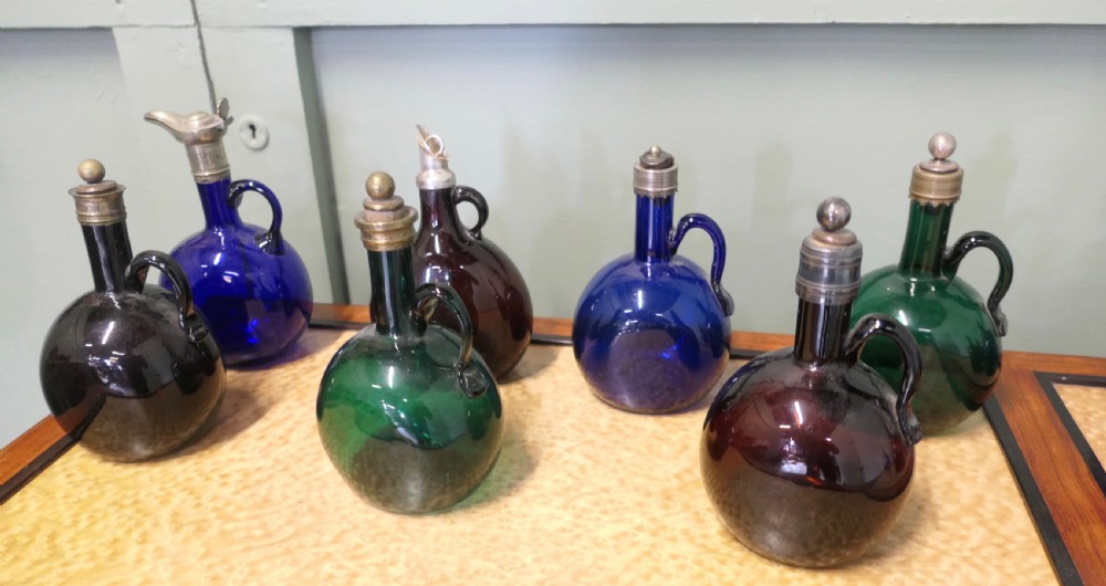 A collection of fifteen 19th-century Victorian coloured glass flagon decanters with metal spouts and stoppers arranged on a wooden surface. View V