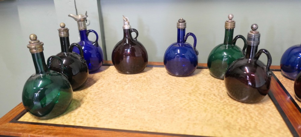 A collection of fifteen 19th-century Victorian coloured glass flagon decanters with metal spouts and stoppers arranged on a wooden surface. View VII