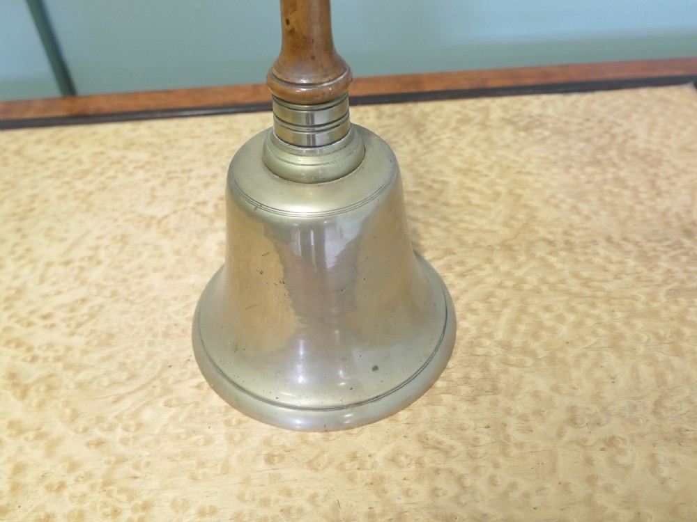 An antique brass hand bell featuring a flared metal skirt, a turned wooden handle, and a brass finial top. View V
