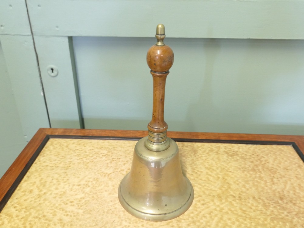 An antique brass hand bell featuring a flared metal skirt, a turned wooden handle, and a brass finial top. View VII