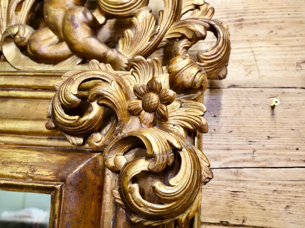 A 19th-century Regency carved giltwood wall mirror featuring a rectangular frame topped with a cresting of two cherubs flanking a flower basket. View V