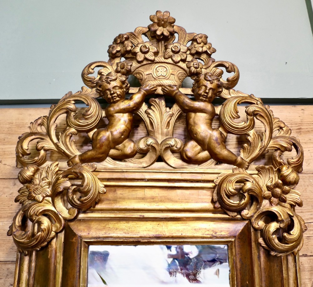 A 19th-century Regency carved giltwood wall mirror featuring a rectangular frame topped with a cresting of two cherubs flanking a flower basket. View VI