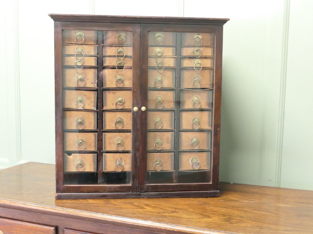 A dark wood glazed haberdashery cabinet featuring twin glass doors enclosing twenty-eight small drawers with brass ring pulls.