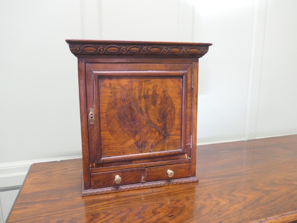 A small burr walnut cabinet featuring a single door with a central circular veneer inlay positioned above two lower drawers with round brass knobs.