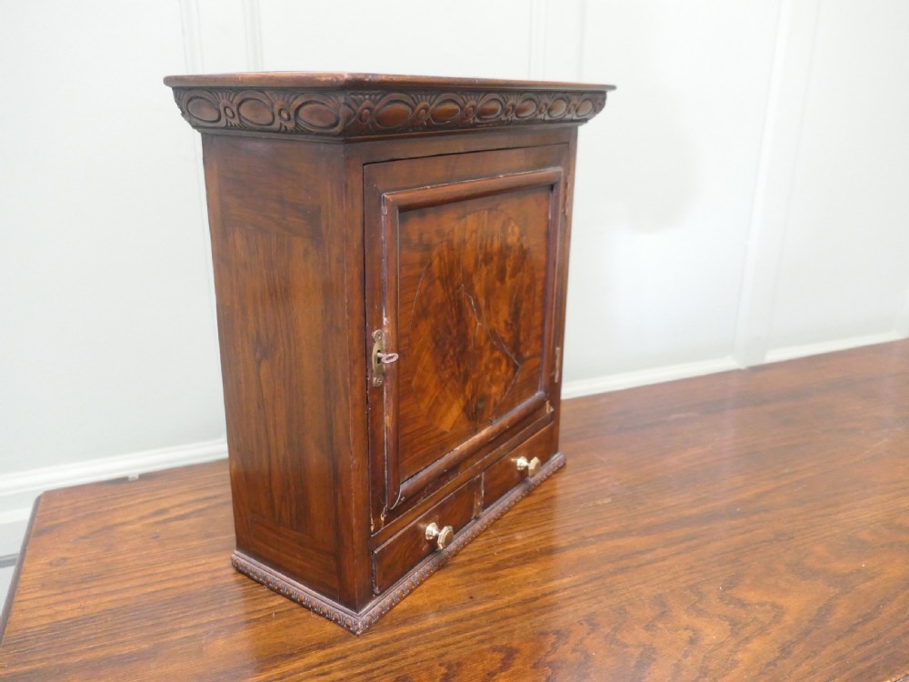 A small burr walnut cabinet featuring a single door with a central circular veneer inlay positioned above two lower drawers with round brass knobs. View II