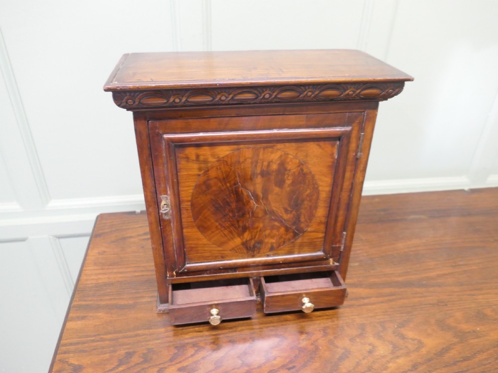A small burr walnut cabinet featuring a single door with a central circular veneer inlay positioned above two lower drawers with round brass knobs. View III