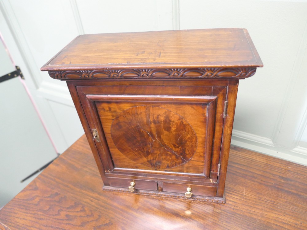 A small burr walnut cabinet featuring a single door with a central circular veneer inlay positioned above two lower drawers with round brass knobs. View V