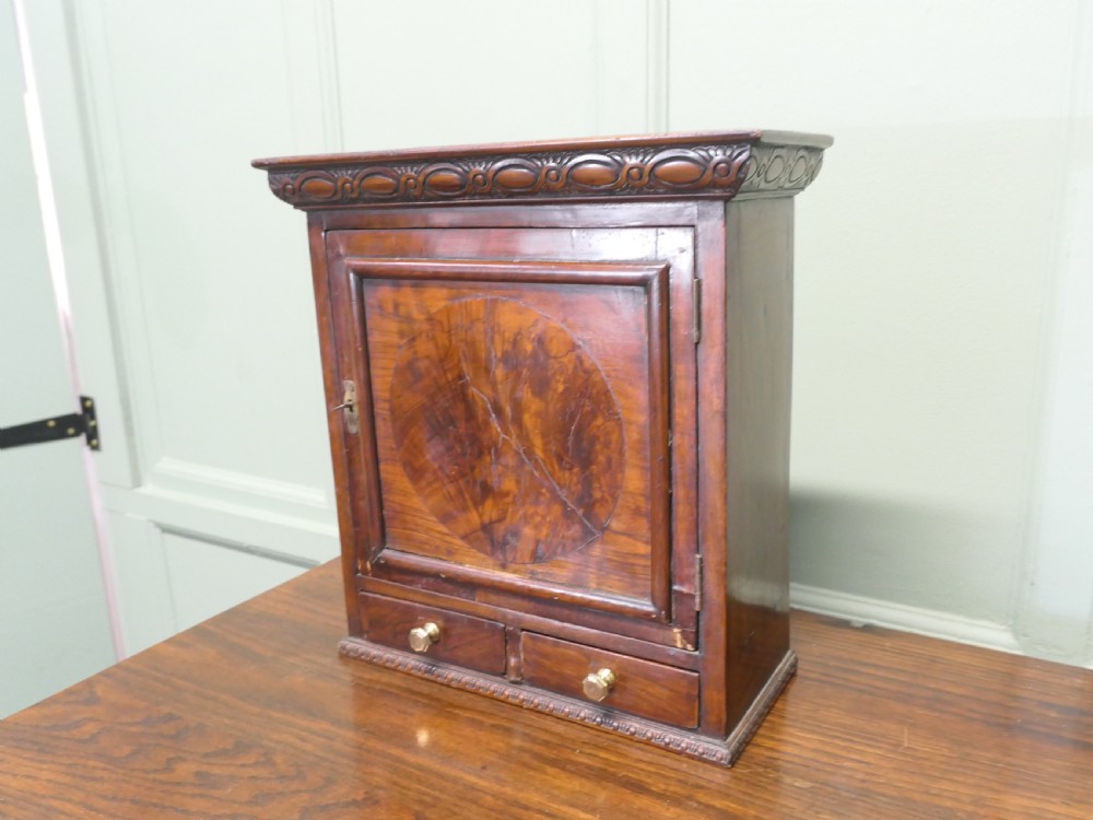 A small burr walnut cabinet featuring a single door with a central circular veneer inlay positioned above two lower drawers with round brass knobs. View VI