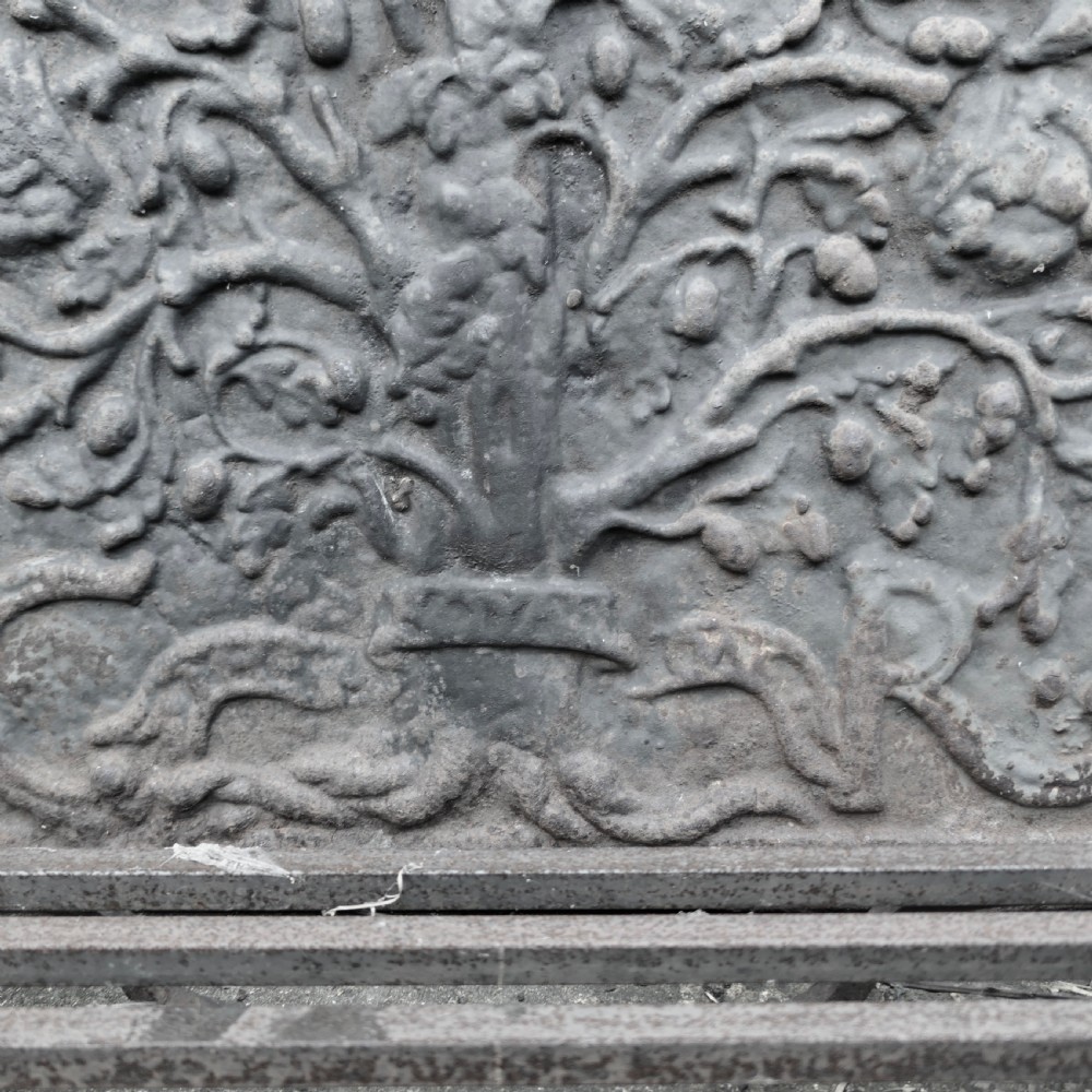 A heavy dark cast iron fireback featuring raised botanical relief decoration positioned behind a large iron slotted fire grate and a pair of tall wrought iron andirons with cresset tops. View IV